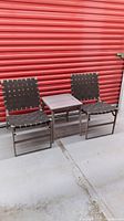 Two outdoor chairs with brown woven fabric seats and backs and metal frames, positioned with a square metal end table with wood-look top between them, in front of a red corrugated metal door.