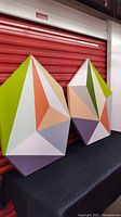 Pair of large polygon-shaped colorful geometric paintings with multi-color sections resting on a table in front of a red storage door.