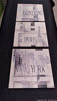 Three city-themed printed canvases laid out on black quilted surface: London, Paris, New York.