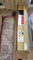 Box of Billie Jean King's Eye Coach tennis training system shown beside a rolled rug and cardboard box.