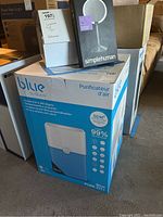Box of Blueair PURE 211+ air purifier in blue and white with product photo and branding, displayed with other boxed items.