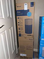 Two boxed Blueair Blue Pure 311 Auto air purifiers stacked upright in corner near a door, showing front and back of packaging with product image and details.