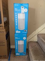 Two boxed Blueair Blue PURE 411+ air purifiers stacked beside a staircase, showing packaging and product branding.