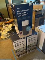 Stacked boxes of three Blueair Blue Pure 411 air purifiers on floor, surrounded by household items.