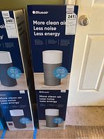 Two boxed Blueair Blue Pure 411 Auto air purifiers next to a door, showing the front and side of the boxes with product branding and information.