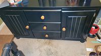 Full view of black wooden buffet showing three drawers with wooden knobs and two cabinet doors with similar knobs, positioned on a floor with visible scratches on the surface.