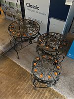 Three different sized metal plant stands with detailed scrollwork and orange faux gems on top