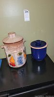 Two ceramic containers on black surface: larger beige Rumtopf crock with fruit designs and smaller plain blue canister with wooden lid