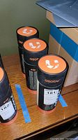 Three Asobu Wireless Beat Bottle packaging tubes standing upright on a wooden surface, showing branded tops.