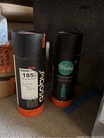 Two boxed Asobu Wireless Beat Bottles with Vacuum Insulation and Bluetooth Speaker, packaged in black cylindrical containers with orange base and turquoise branding.