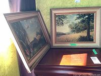 Two framed landscape paintings placed on a wooden cabinet, showing the overall condition and arrangement.