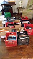 Eleven boxes/crates filled with books arranged on a wooden floor in a living room space. Crates are plastic and of various colors including red, black, and green. Books are packed vertically and horizontally inside.