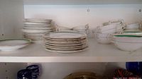 Shelf view showing stacks of plates, cream soup bowls with handles, and teacups with handles on white china with green and gold trim.