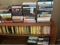 Books arranged on wooden headboard shelf and stacked on top, viewed frontally showing varied titles and conditions.