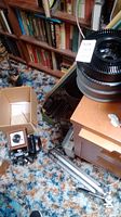 Photo showing the projector with slide trays on top and the vintage camera on carpeted floor near bookshelf.