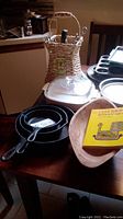 Photo of cast iron pans, clay baking dish, Corningware microwave fryer with lid, woven basket with utensils and a yellow book 'Clay Baking'.