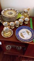 Full lot display showing eight cups with gold handles, eight saucers stacked, side plates with romantic scenes, and one blue bordered serving plate.