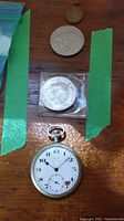 Wooden surface with four items: three coins and one pocket watch. One coin is in a plastic protective square, another positioned on top of a larger coin, and pocket watch below them.