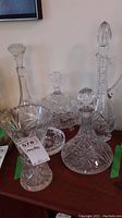 Six clear crystal items on a wooden surface, including three decanters with stoppers, cut crystal vase, covered bowl, and ashtray.