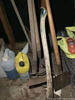 Photo showing various wooden-handled garden tools including shovel, pitchfork, and hoe arranged against a wall along with two plastic fluid containers on the floor.