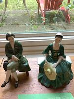 Front view of two Royal Doulton Williamsburg figurines seated by a window on a wooden surface.