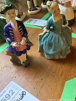 Two Royal Doulton colonial Williamsburg mini porcelain figurines including a boy holding a musket and a girl in a blue dress, shown from the front on a wooden surface