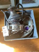 Photo showing a crate filled with silver plate hostess ware including a coffee pot, several serving trays, a warming stand, and a small dish with a purple liner. Items show wear and tarnish.