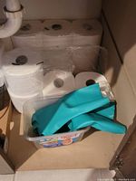 Packages of multiple toilet paper rolls and blue rubber gloves in a container inside bathroom cabinet.