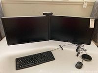 Two Dell monitors mounted on a dual desk stand with nearby Logitech keyboard, wired Tecknet mouse, and round Tonor microphone speaker on a white surface.