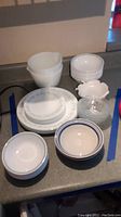 Sixteen Corelle white bowls with blue stripe, fourteen matching plates, two white Pyrex mixing bowls, and four small glass bowls arranged on countertop.