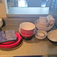 Photo of various stacked dishware including red bowls, pink plates, white mugs, and stacked white serving bowls on a countertop.