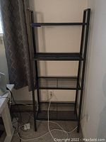 IKEA Lerberg metal shelving unit in dark grey, showing four slatted shelves against a wall near a curtain and power cords.