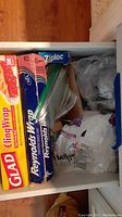 Photo showing kitchen drawer with Glad Cling Wrap, Reynolds aluminum foil, Ziplock baggies, and paper napkins stacked inside.