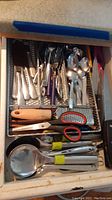 Top-down view of drawer with various silver cutlery, kitchen scissors, and a potato peeler.