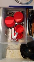 Top-down photo of translucent plastic bin containing kitchen utensils including red measuring cups and spoons, metal can opener with white handles, whisk, ladle, and other metal utensils.