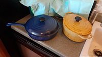 Photo showing blue Le Creuset skillet and yellow Dutch oven side by side on countertop.