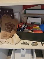 Photo of home office supplies including scissors, envelopes, paper bags, and other stationery items placed on a white shelf with some small decorative magnets visible below.