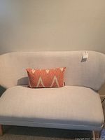 Front view of the light-colored linen loveseat sofa with an orange and white geometric patterned decorative pillow on seat.