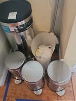 All waste baskets and trash cans together in corner; three small stainless steel cans, one large stainless steel can with black lid, and one beige plastic waste basket.
