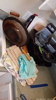 Photo showing large metal wok with wooden handle, metal box grater, small metal strainer, wooden cutting board, metal baking sheets, and stacked fabric napkins and placemats.