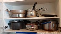 Shelf with assorted pots and pans including stockpot, saucepans with lids, frying pans and a colander.