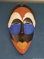 Front view of the painted African mask, prominently displaying red-orange, black, white, blue, and yellow ochre colors and facial features.