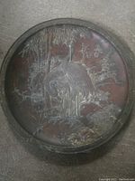 Front view of antique Asian bronze plate showing raised design of crane standing among bamboo and other plants.