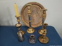 Photo shows six candlesticks and one decorative brass tray arranged on a blue surface against a white wall. Tallest brass candlestick has a candle.