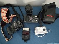 Lot photo showing Minolta 7000 camera with lens, Minolta Maxxum flash, Kodak Easy Share camera, Pentax Zoom 90 WR camera with case, and an extra Minolta lens on blue background.