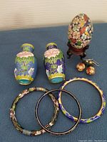 Image shows two small cloisonne vases with colorful floral designs, a cloisonne egg on a wooden stand with a butterfly and flowers pattern, a pair of spherical cloisonne earrings, and three cloisonne bangles with floral motifs.