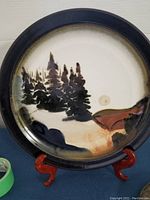 Photo showing a round display charger with a hand-painted hillside scene featuring evergreen trees and sun, bordered by a dark rim, placed on a wooden stand.