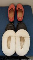 Pair of reddish Clark's women's shoes on top and black Garneau men's sheepskin slippers with white lining below, both pairs displayed on dark surface