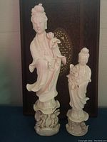 Two white porcelain Quan Yin Blanc de Chine figurines, one larger and one smaller, posed with flowing robes and lotus flowers, set against a decorative backdrop.