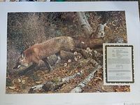 Photo of the art print featuring a red fox walking in an autumnal forest scene with a certificate of authenticity placed on the print.